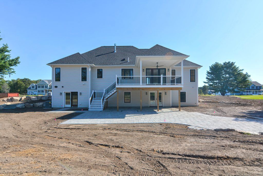 Rear view of a light gray house with deck and glass railings under a clear blue sky on a dirt lot.