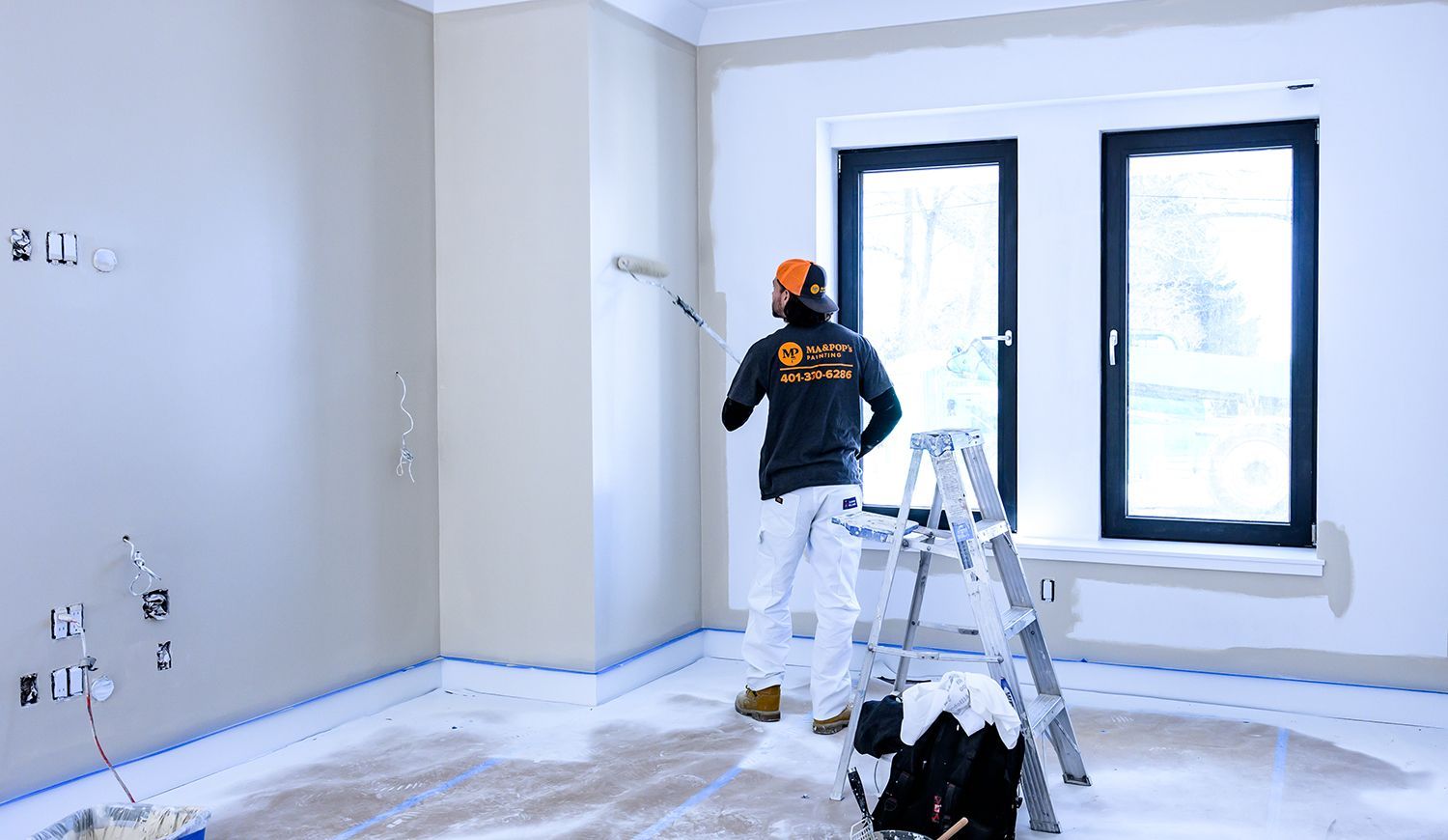 Painter in a room, using a roller on a wall. Ladder and masking tape visible.