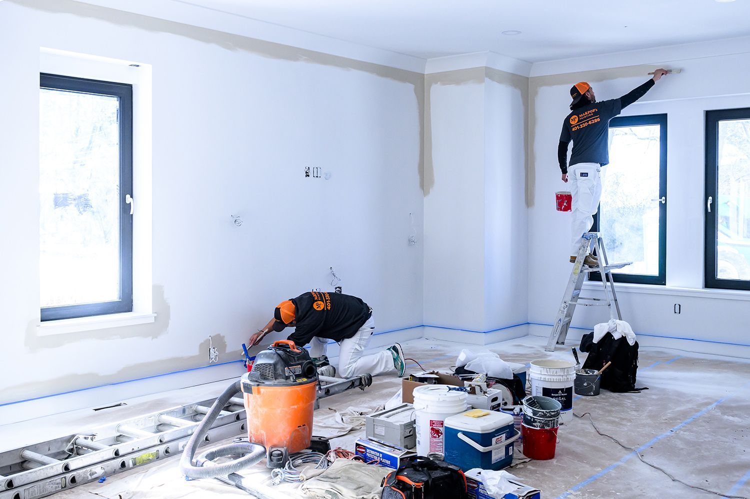 Two people painting a room. One on a ladder, the other near the floor, both wearing work clothes and orange hats.