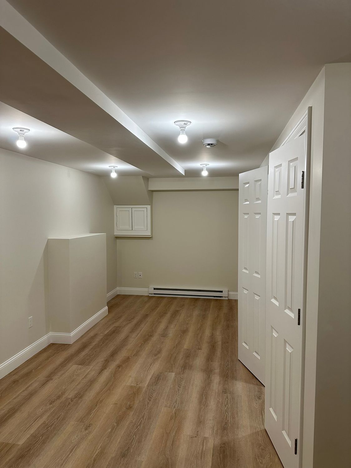 A finished basement with wood-look flooring, white walls, recessed lights, and closed white doors.