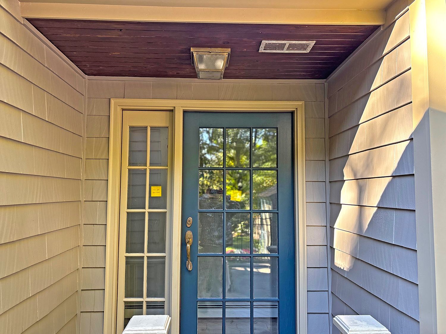 Blue door and sidelight under a porch, light streaming from the right, gray siding, brown ceiling.
