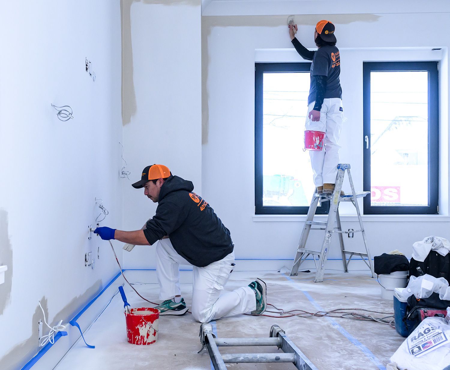 Two people painting a room. One on a ladder, one kneeling. White walls, windows, and equipment present.