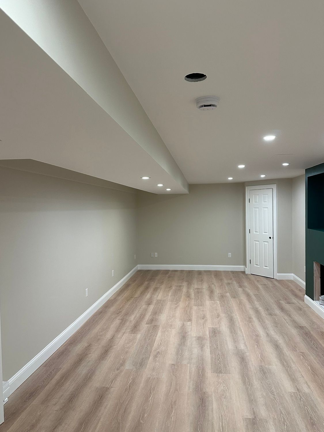 Empty, light-filled basement room with wood-look flooring, recessed lighting, and a white door.