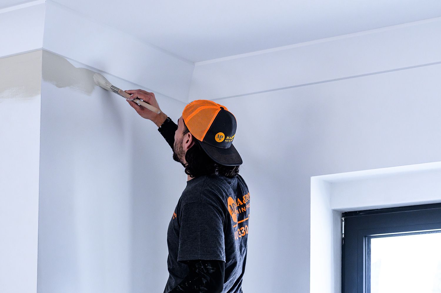 Person painting a white wall near a ceiling, wearing a black and orange hat and a dark gray shirt.