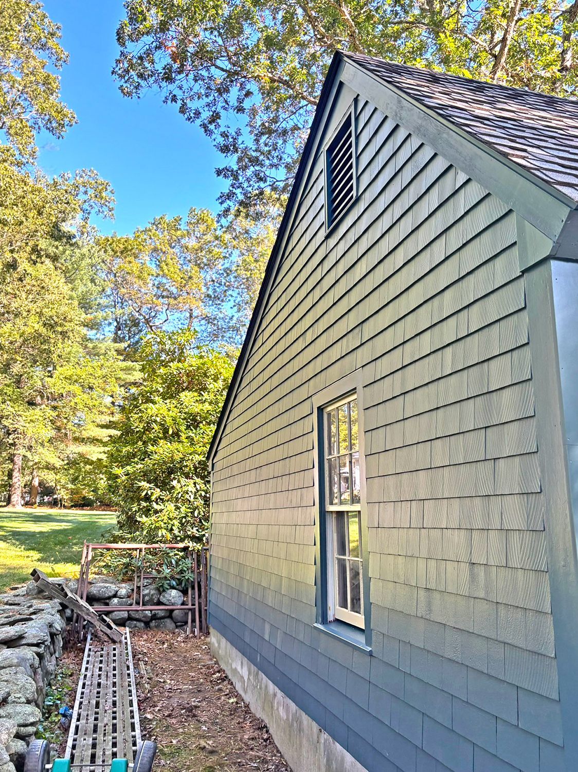 Blue-gray shingled building with a window and a vent, set against a backdrop of trees and a blue sky.