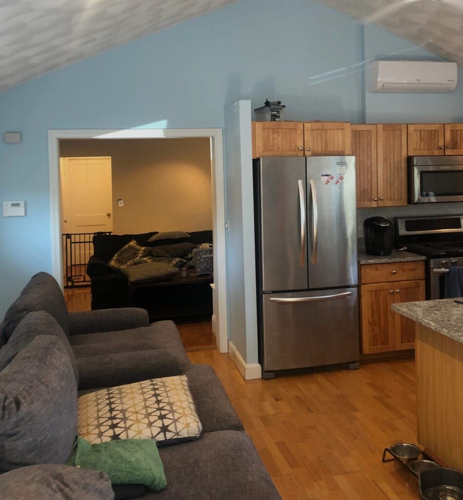 A kitchen and living room interior. Stainless steel fridge, wooden cabinets, and a blue wall. Couch and doorway to another room.