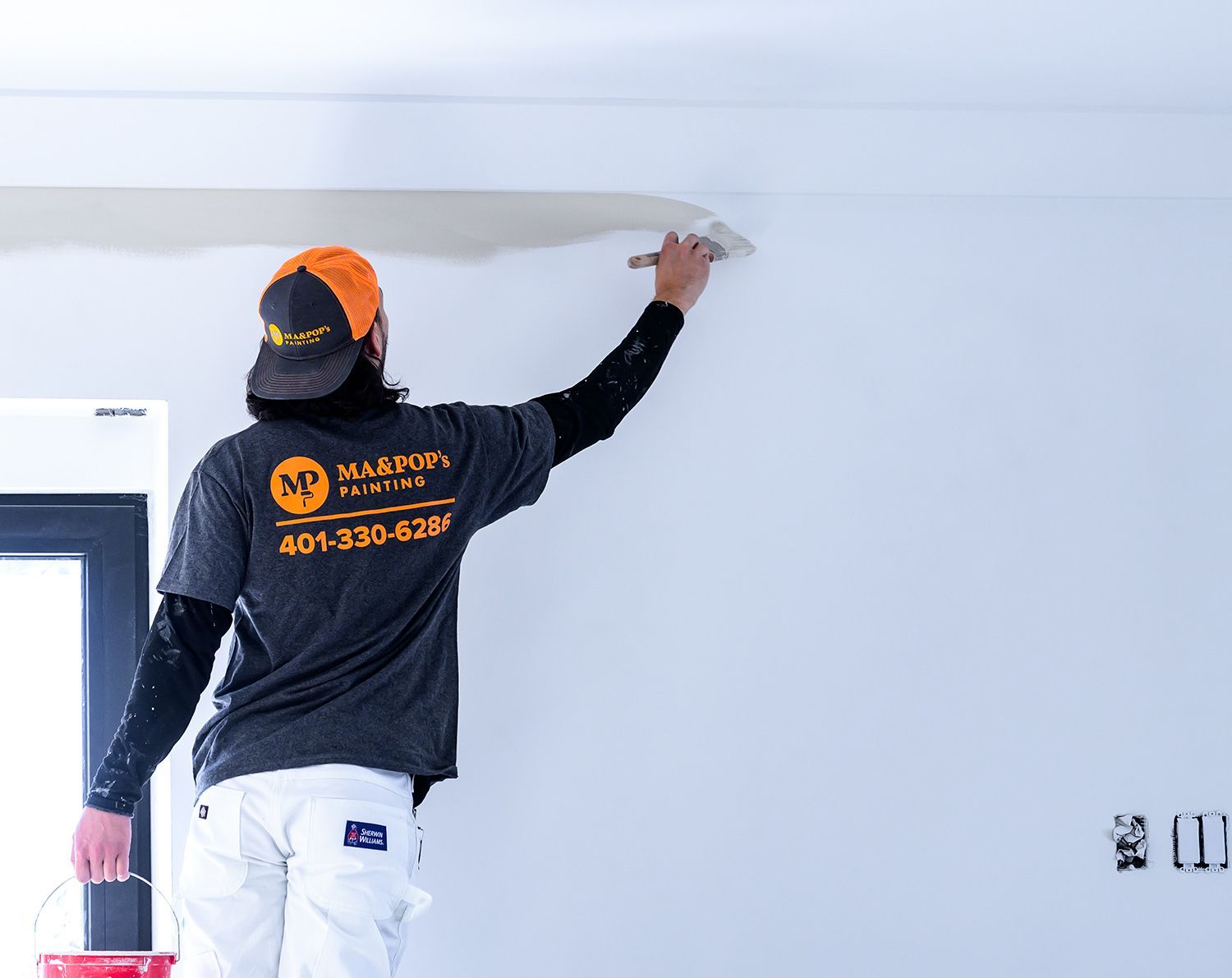 Person on a stepstool applying spackle to a wall's edge. Wearing a work shirt and hat, working in a room.