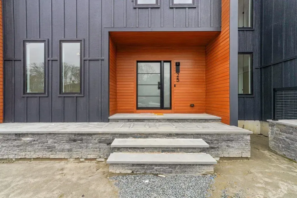 Modern home exterior with gray siding, orange accent wall, and stone steps leading to a glass door.
