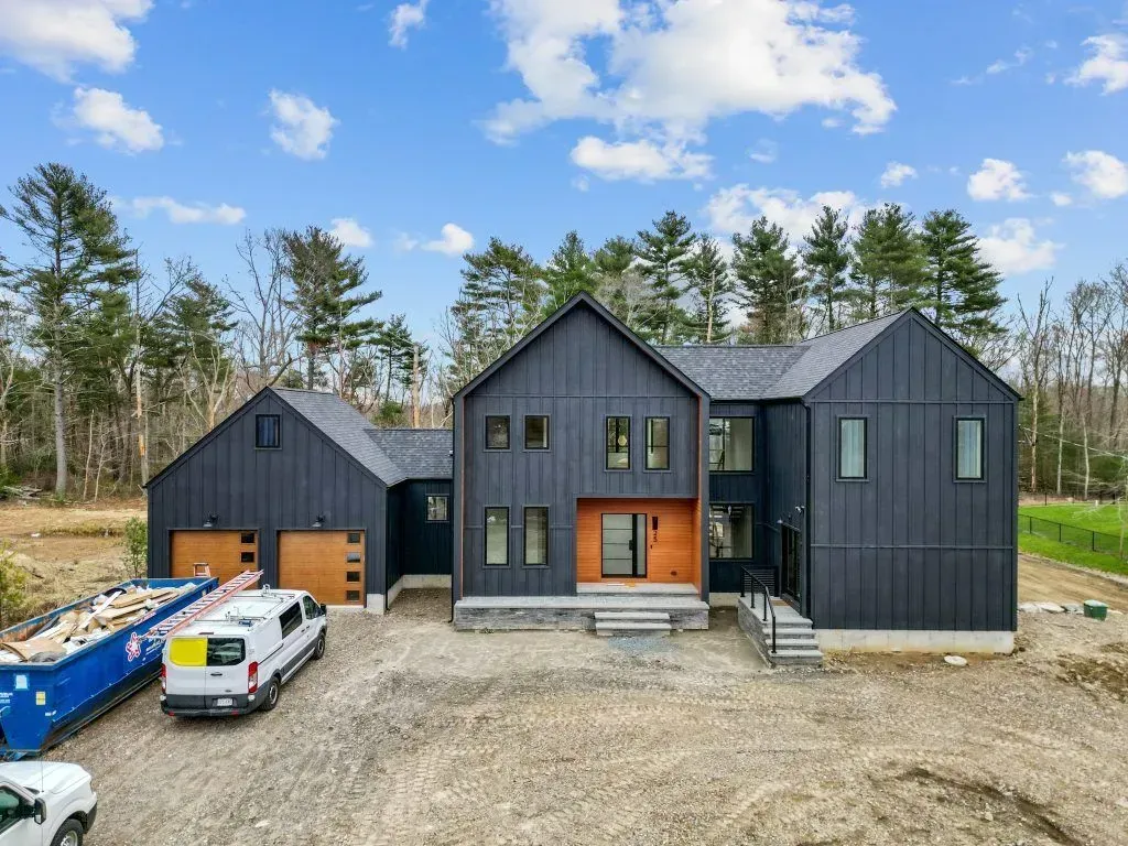 Modern black house with brown garage doors, standing on a dirt lot. A blue dumpster and a white van are in front.