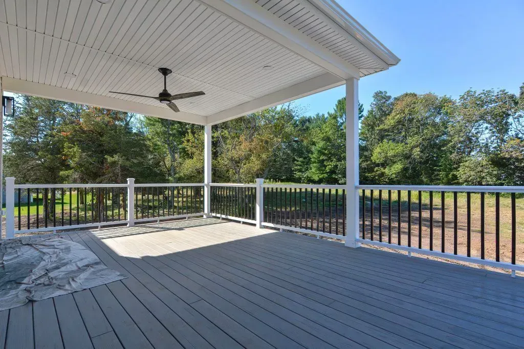 Covered deck with gray flooring and black railing, overlooking a field with trees under a blue sky.