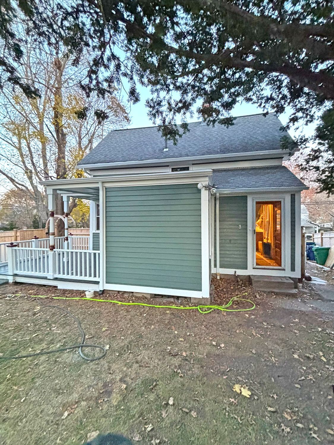 Small teal house with white porch and railing, next to a tree. Door is open, and light is visible.