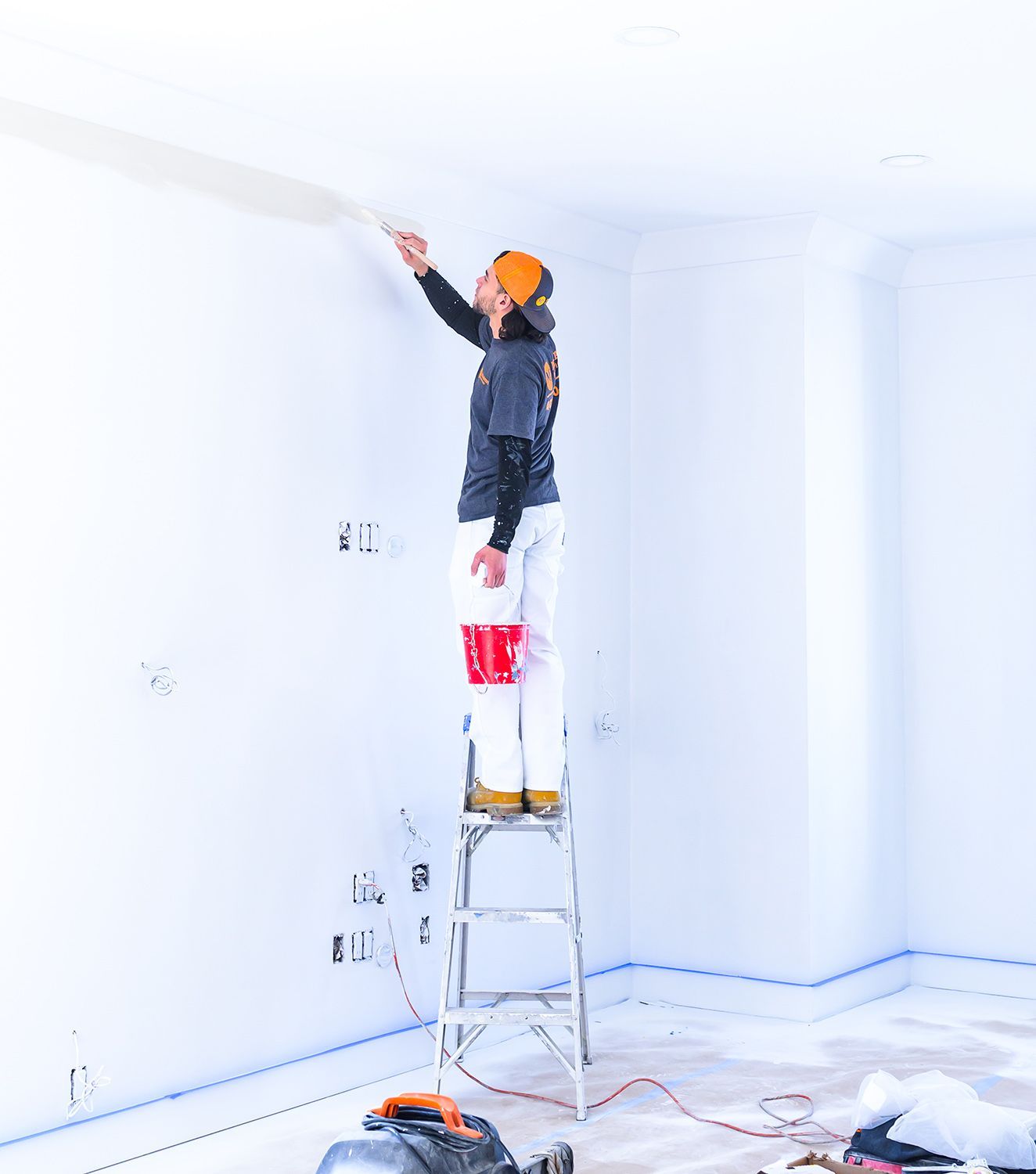 Person painting a white wall, standing on a ladder. Interior of a room with trim, tools on the floor.