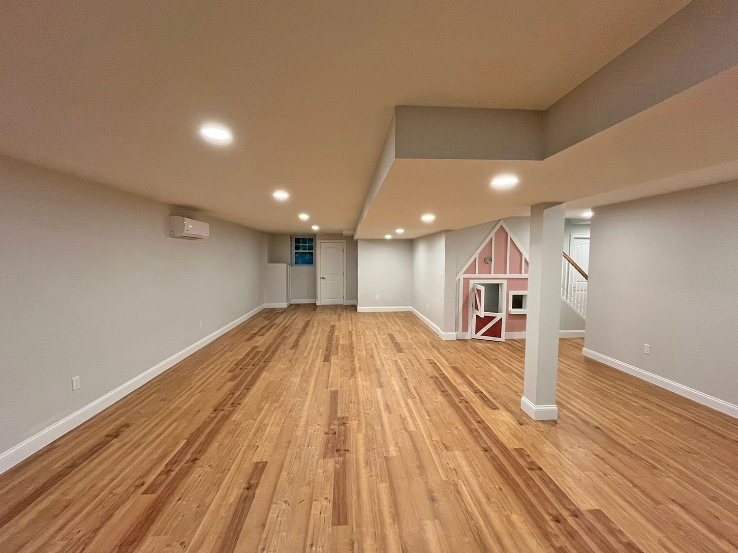 Empty basement room with light wood floors, gray walls, and recessed lighting. A pink playhouse sits in the corner.