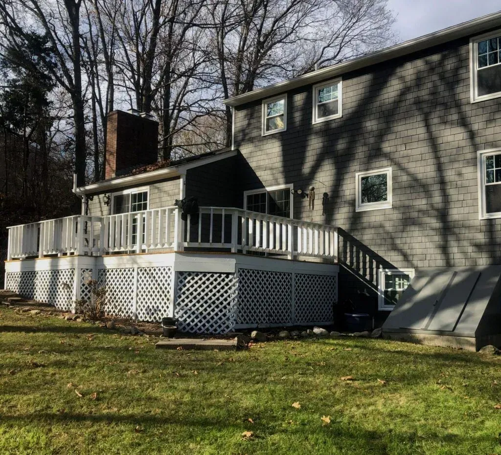 Rear view of a two-story gray house with a large deck, surrounded by a grassy yard.