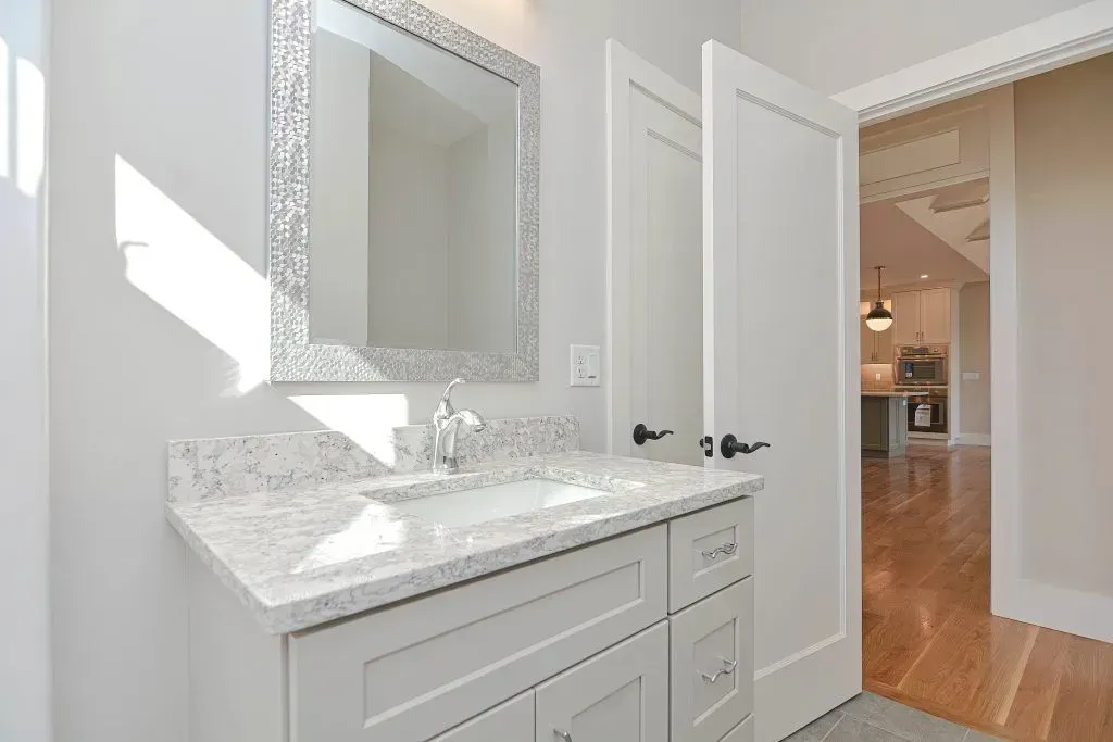 Bathroom with white vanity, speckled countertop, and silver-framed mirror. Open door leads to kitchen.