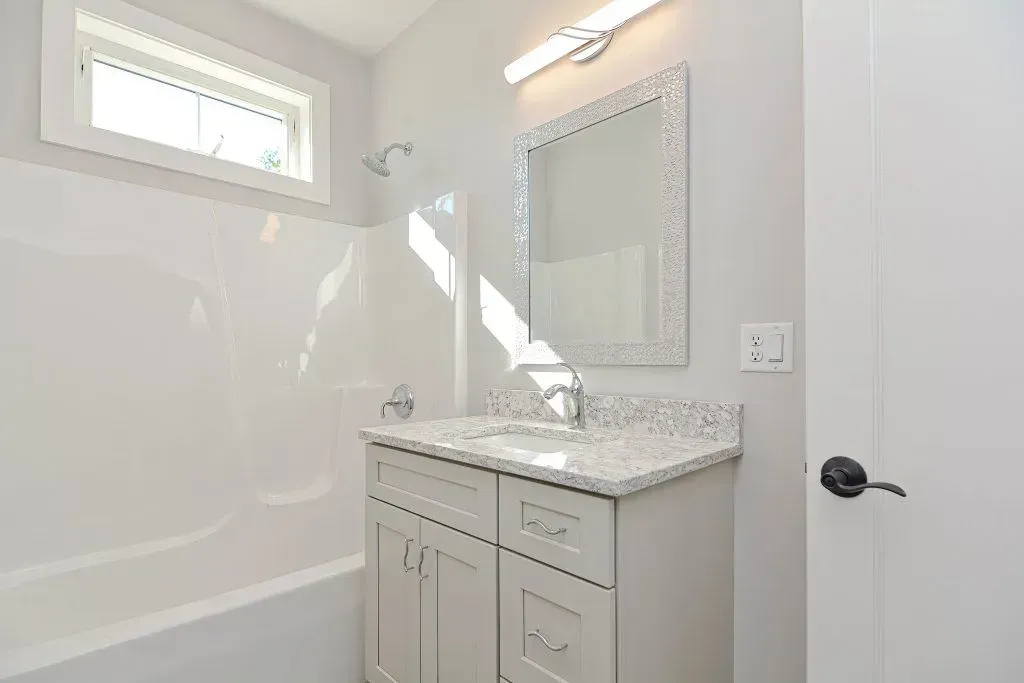 Bathroom with white walls, tub, vanity, and window. Gray countertop, decorative mirror.