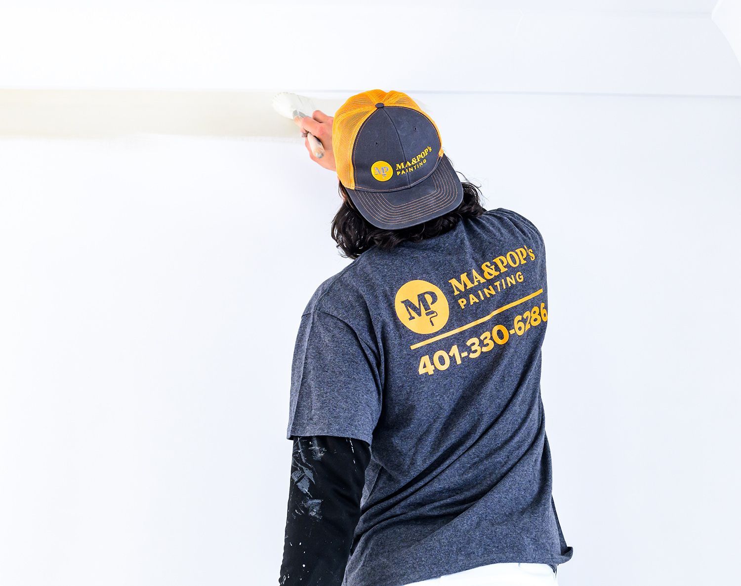 Painter applying white paint to a ceiling trim. The person wears a baseball cap and t-shirt with a painting company logo.