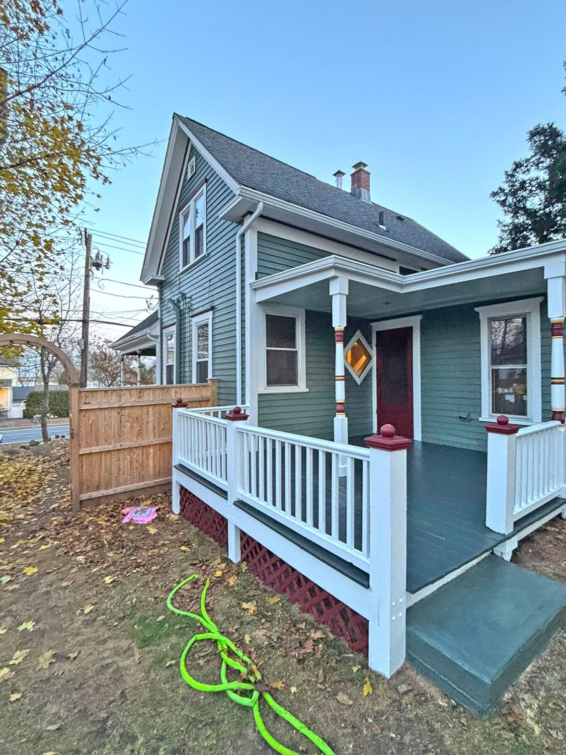 Green house with white porch and railing, red door, and wooden fence on the side.