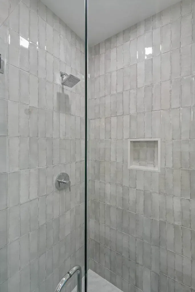 Shower with gray vertical tiles, glass door, and built-in shelf. Silver shower head and faucet.