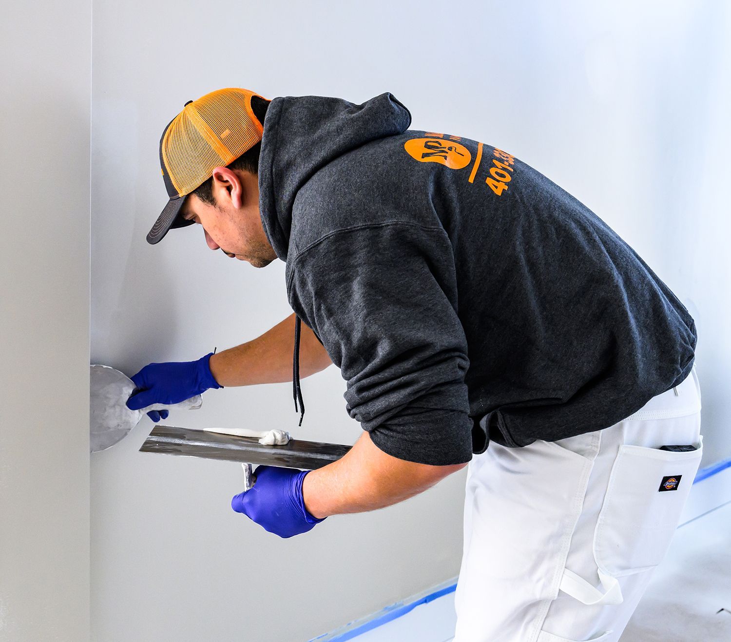 Man in hoodie and cap, applying compound to a wall with a trowel, wearing gloves.