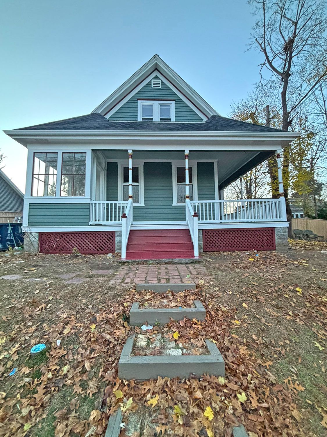 A light teal house with a porch and red trim, set on a brown leaf-covered lawn.