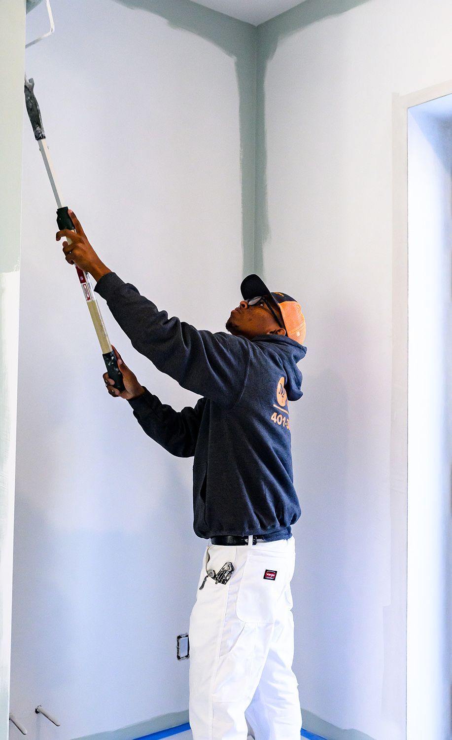 Person painting a corner of a room with a roller, wearing safety gear and paint-stained pants.