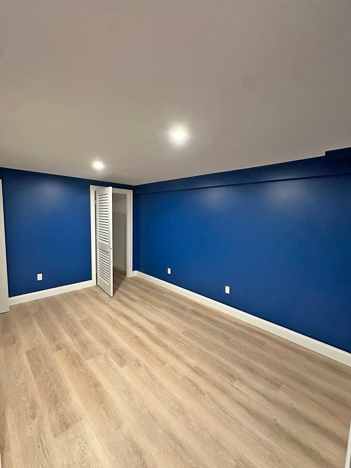 Empty room with blue walls, white trim, recessed lighting, and light wood-look flooring. Closet with open, white door.