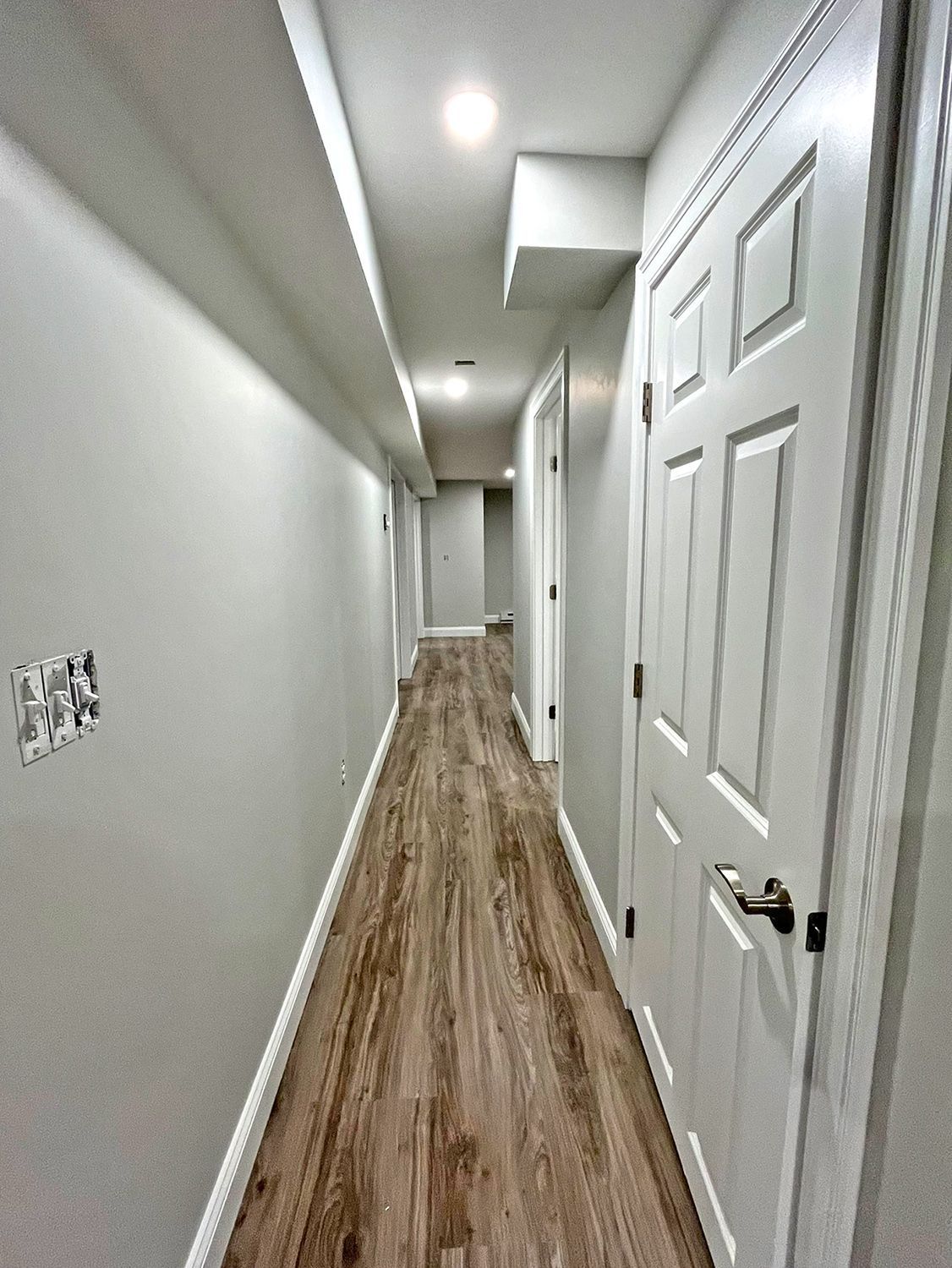 Long, narrow hallway with wood-look flooring, white doors, and light gray walls.