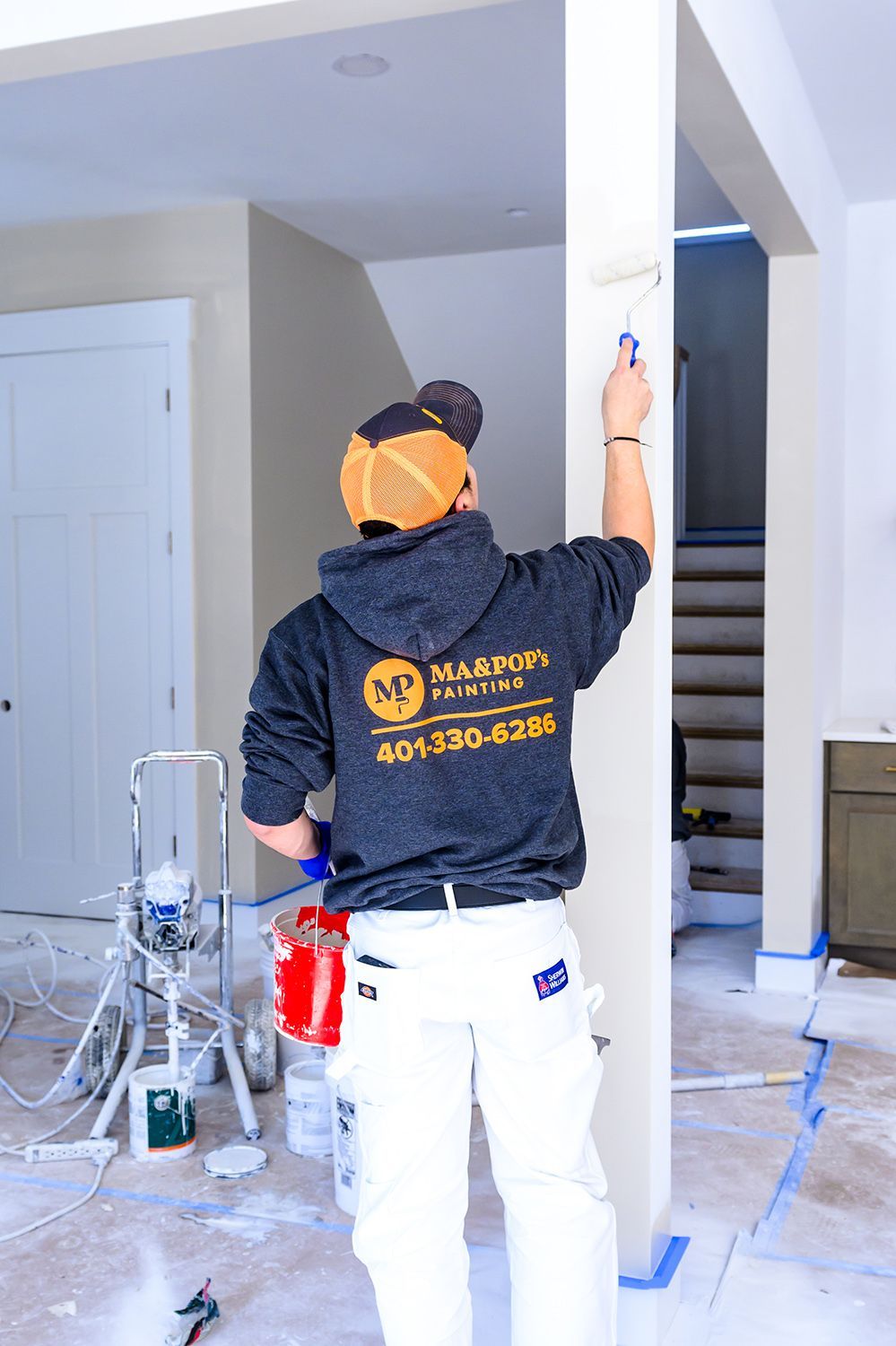 Painter in a hoodie and white pants rolling paint onto a white column in a room under construction.