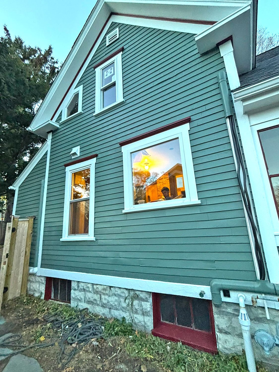 Green house with white trim, red window accents, and stone foundation. Evening lighting reflected in windows.