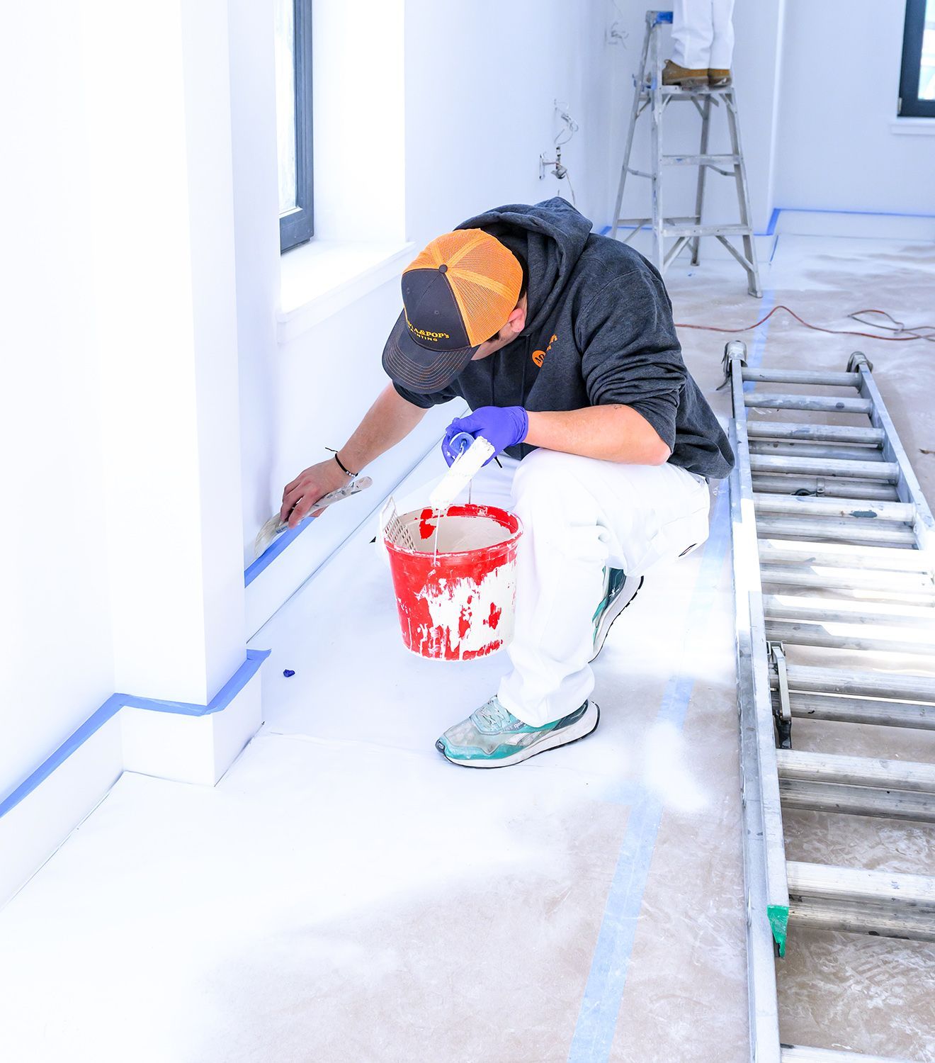 Person painting trim in a room; blue tape, red bucket, ladder visible.