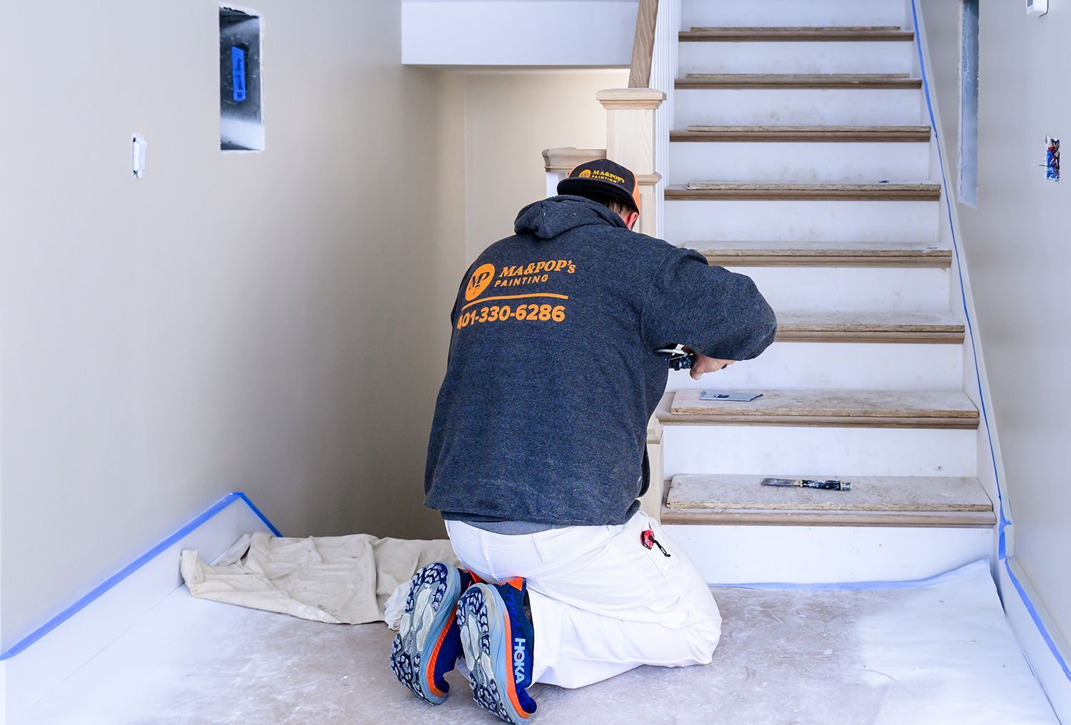 Person kneeling, painting stairs, in a room with blue painter's tape on the floor.