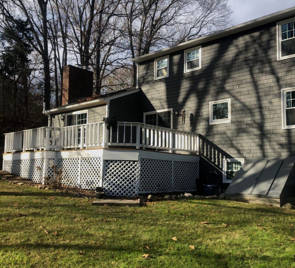 Gray house with white deck, lattice, and stairs, surrounded by trees and a grassy yard.