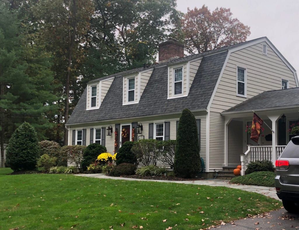 A light-colored house with a dark gray roof. The front yard has a green lawn with shrubs and trees.