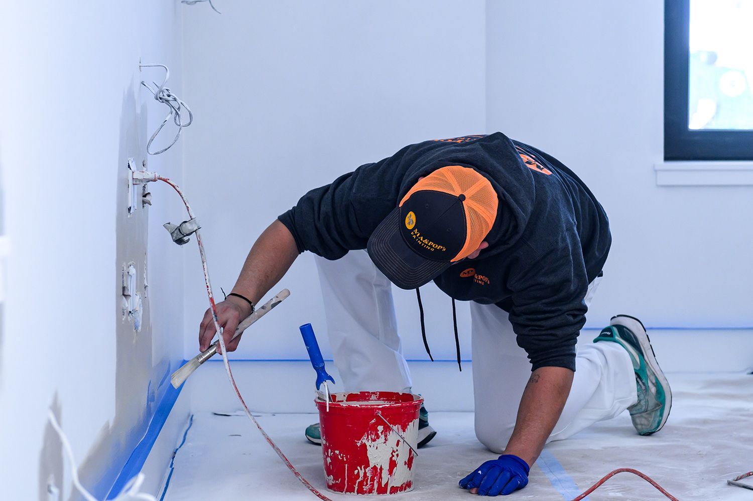 Person paints a wall while kneeling, with a paint roller and bucket. They wear a cap and work gloves.