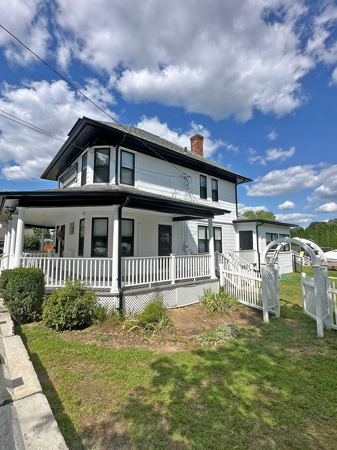 White two-story house with black trim, a wraparound porch, and a white picket fence on a sunny day.