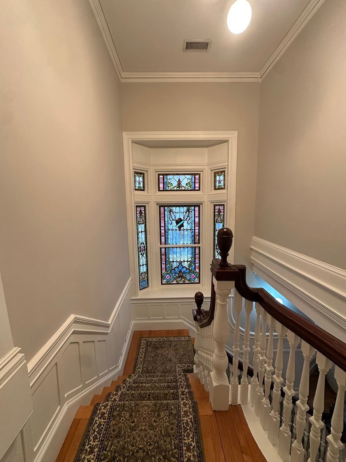Stairway with stained glass window. Cream walls, wood banister with ornate details, patterned carpet runner.