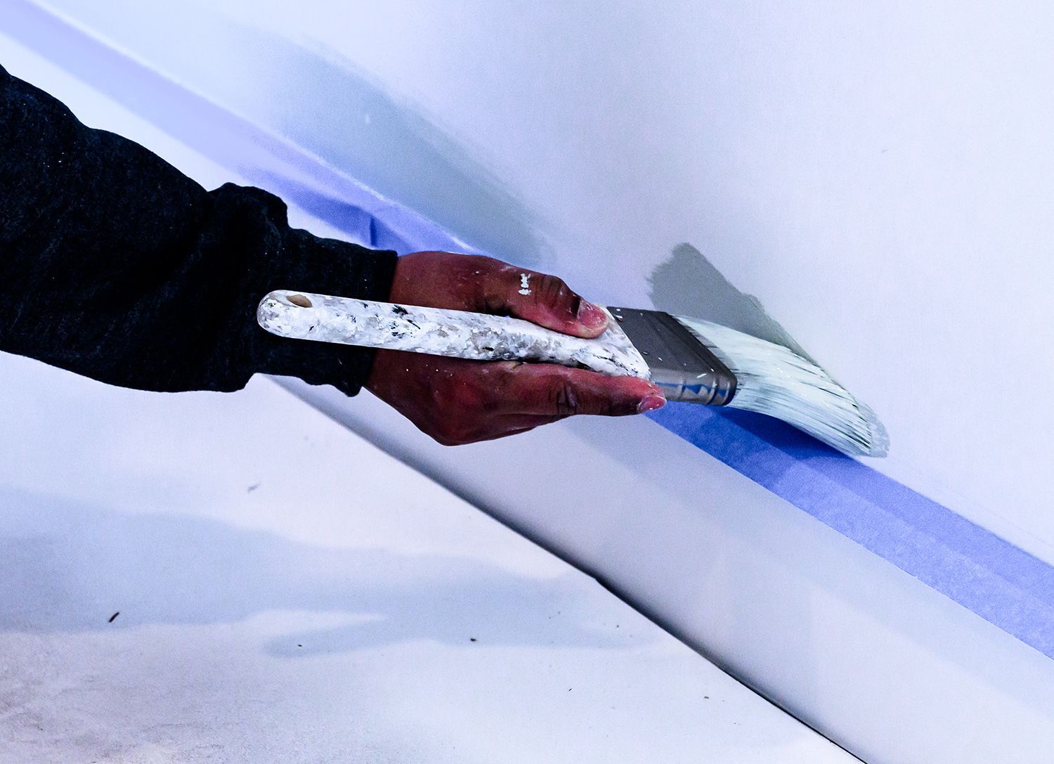 Hand with paintbrush applying white paint to wall, next to blue painter's tape.