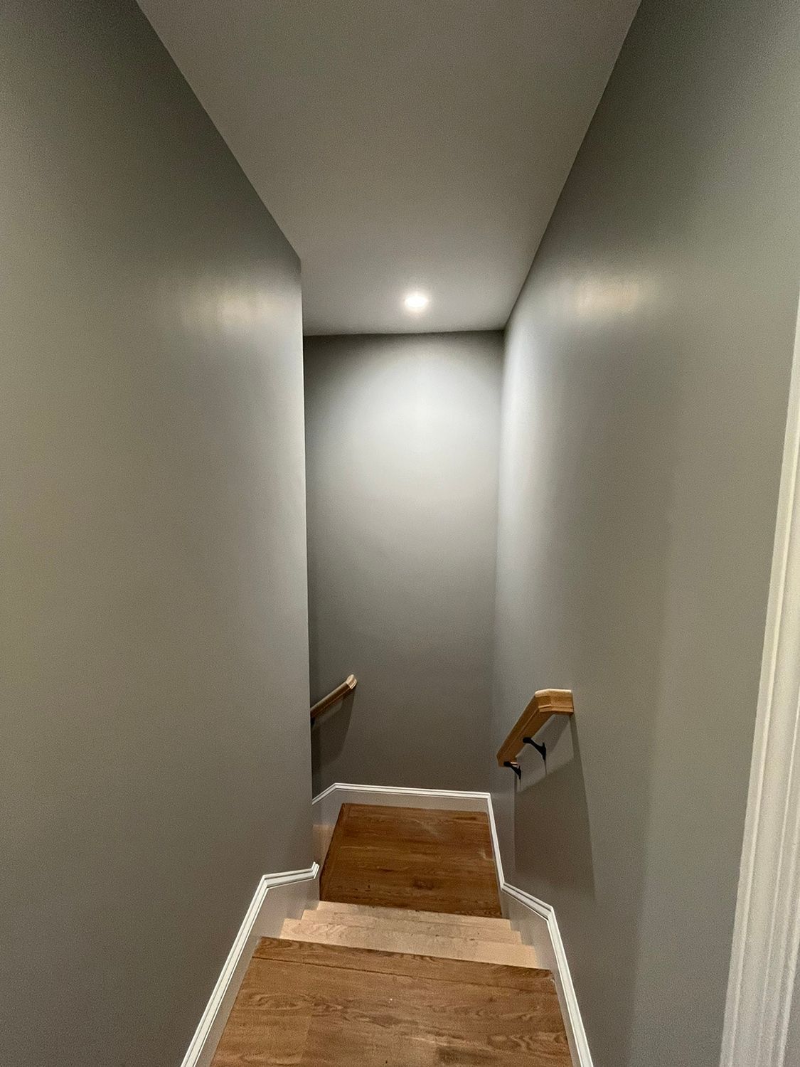Stairwell with gray walls and wooden steps. Handrails on both sides. Overhead light at the end.