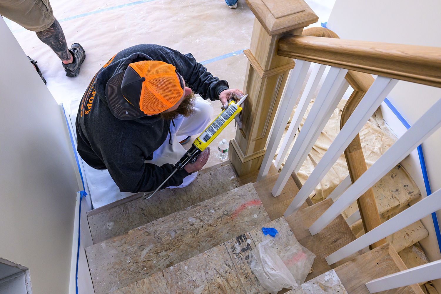 Man using a caulk gun on stairs, applying sealant to a wooden handrail, indoor setting.