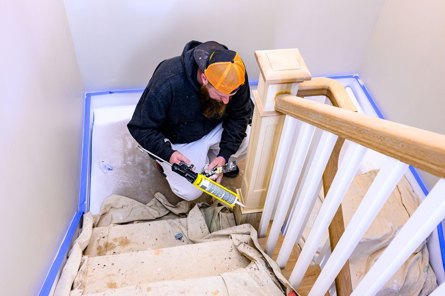 Person caulking stairs, wearing a hooded sweatshirt and orange hat. The steps are covered with drop cloths and blue tape.
