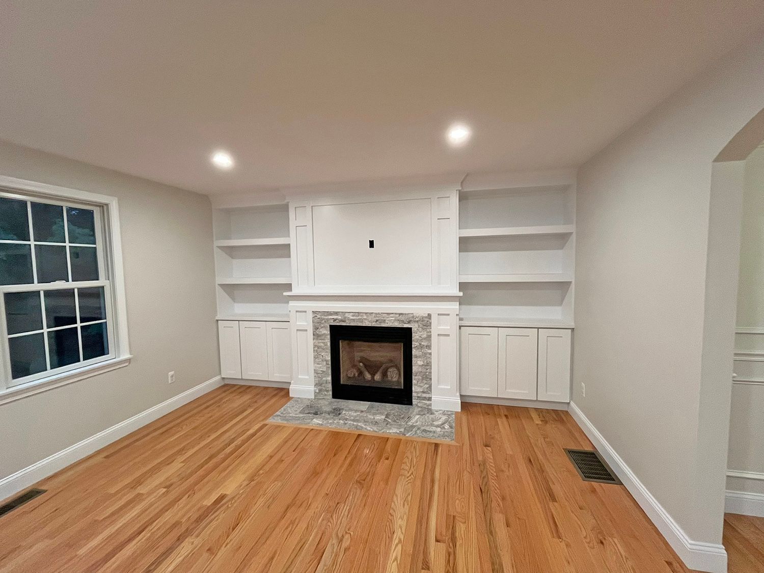 Living room with fireplace, built-in shelves, hardwood floors, and a window. The walls are light grey.