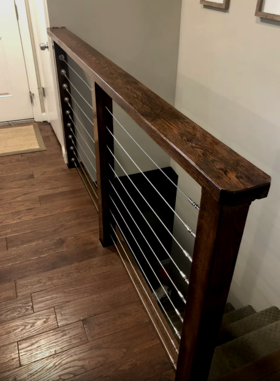 Wooden handrail with stainless steel cables, on stairs. Dark wood and flooring, gray walls.