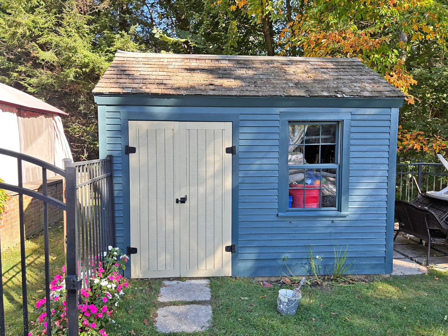 Blue shed with off-white door and small window. Stone path leads to the entrance. Fence and flowers in the foreground.