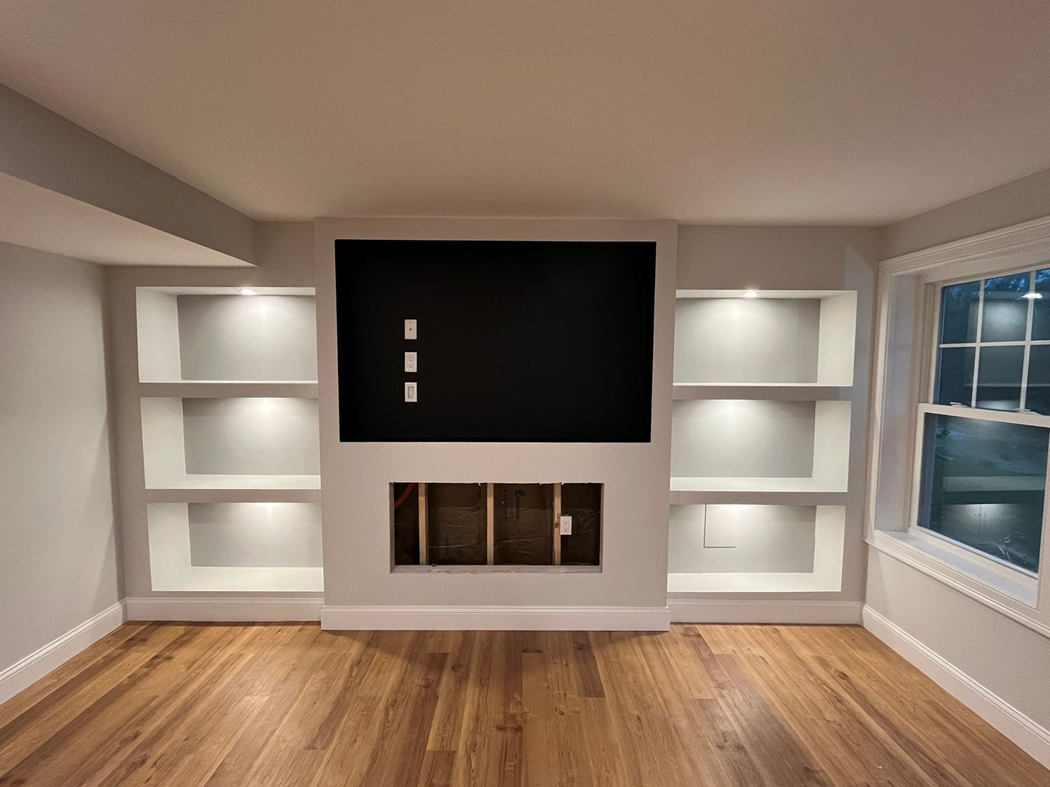 Built-in entertainment center with a black TV, shelves with lights, and a fireplace in a room with hardwood floors.