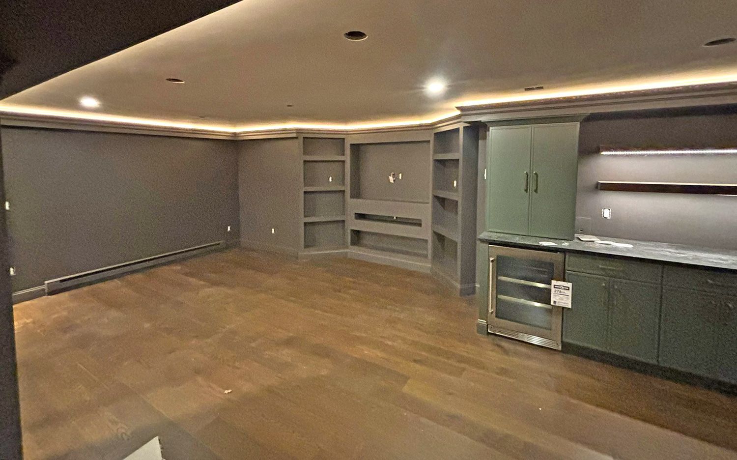 Empty renovated basement with built-in shelves, bar, and wood flooring. Features dark gray walls and ceiling lighting.