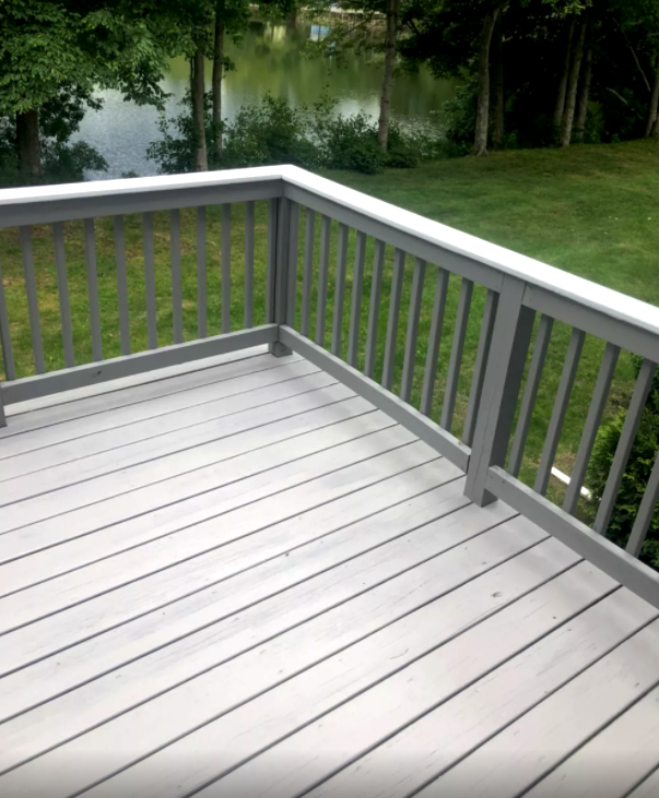 Gray wooden deck with gray railings, white handrail, overlooking a lake.