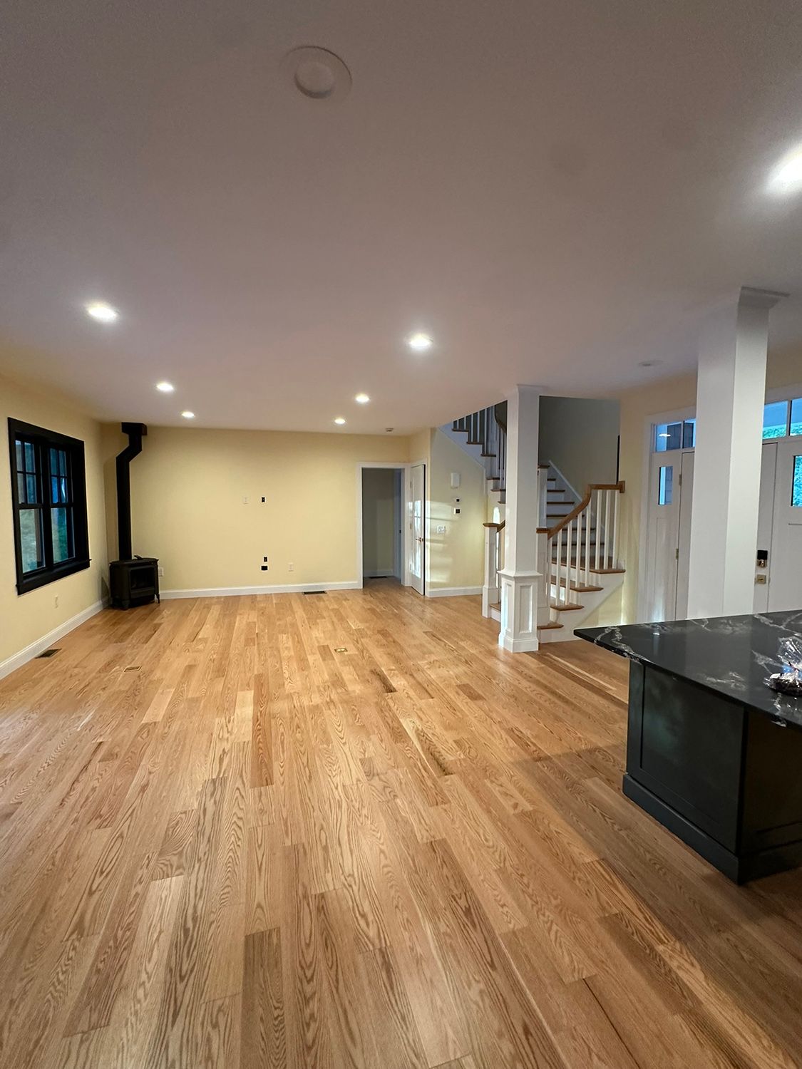 Spacious empty room with hardwood floors, recessed lighting, and a staircase. Fireplace and kitchen island visible.