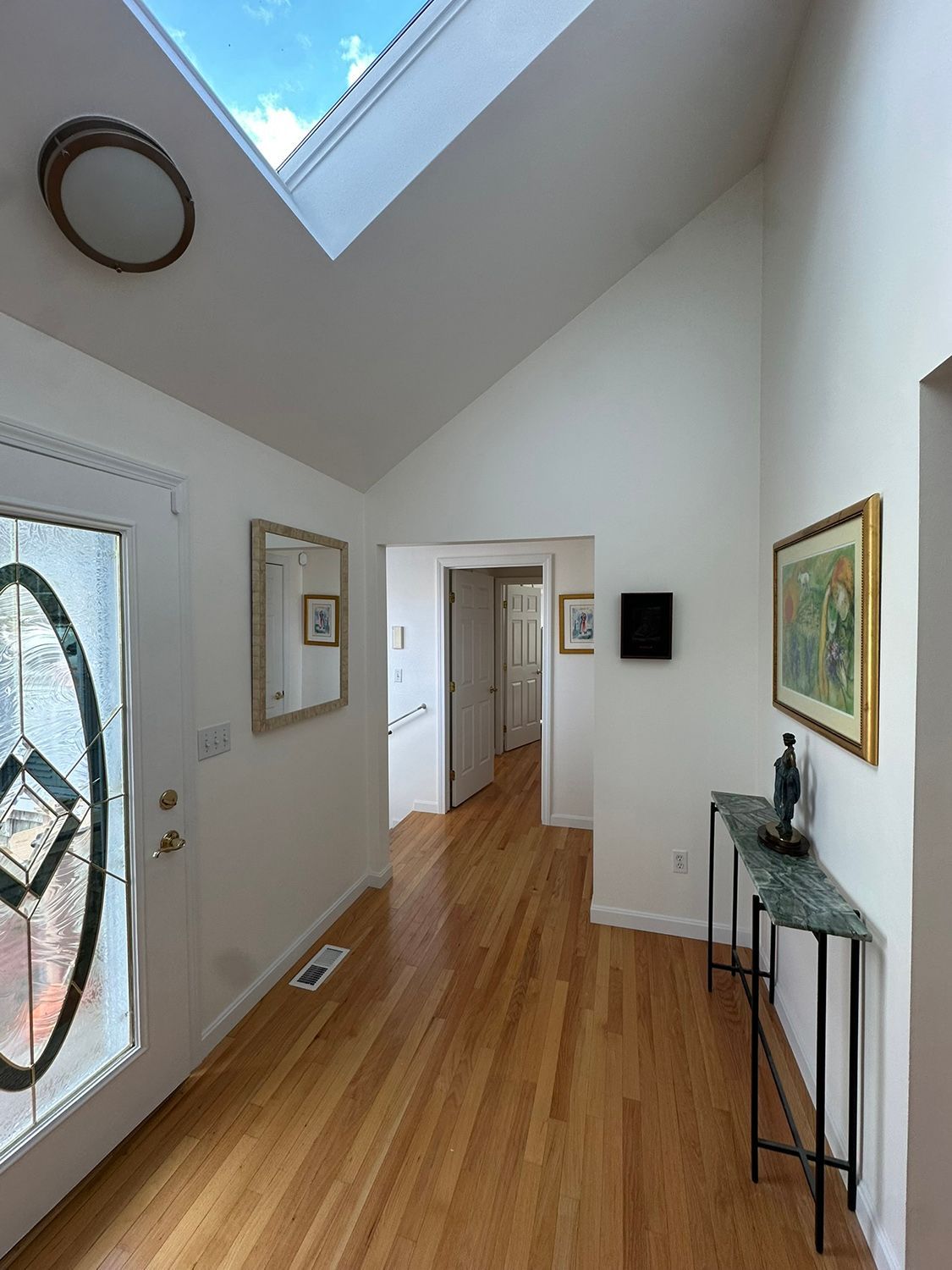 Entryway with hardwood floors, a skylight, and white walls. A table with a marble top, artwork, and a decorative door.