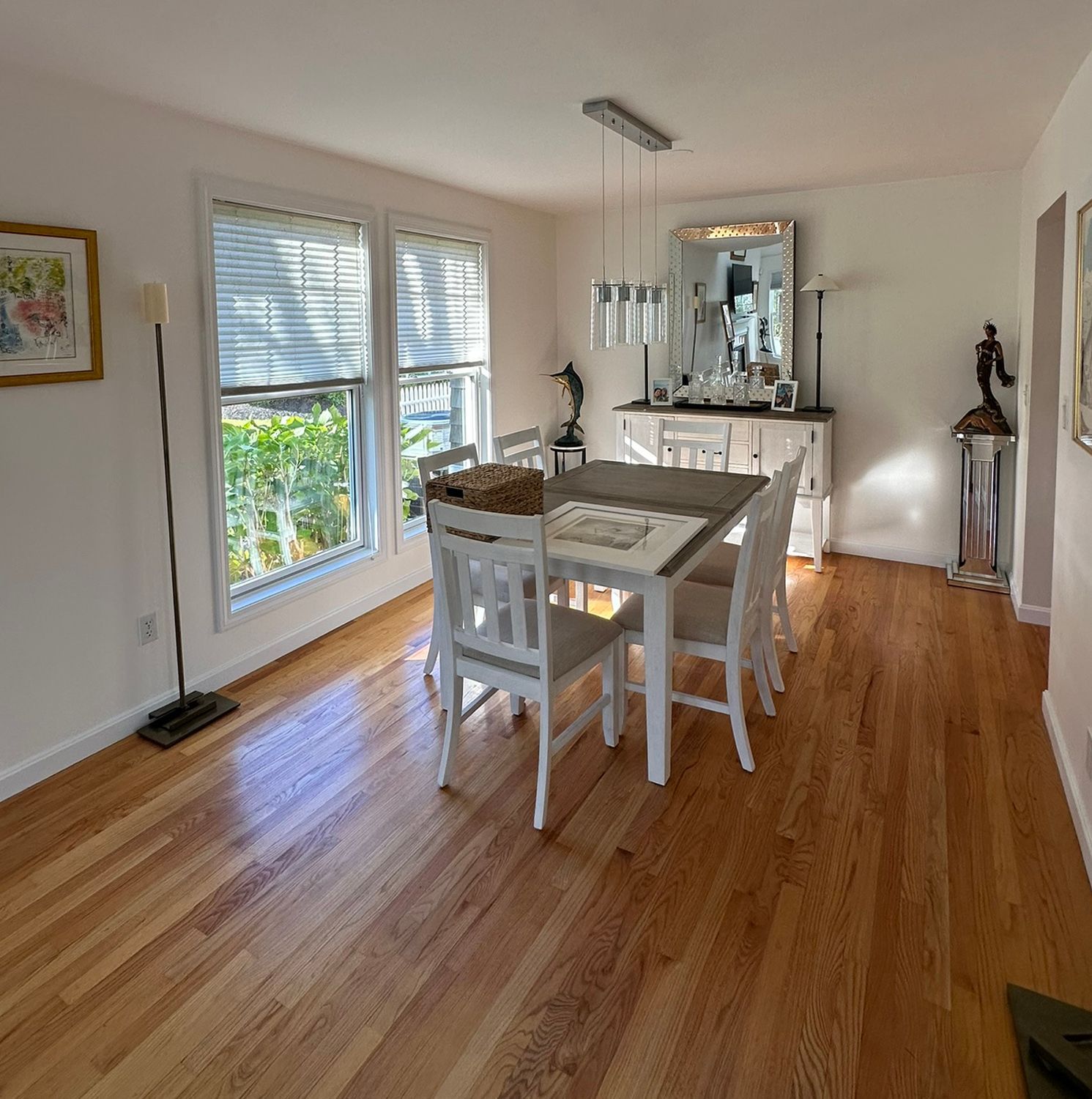 Dining room with hardwood floors, white table, chairs, and buffet. Windows with blinds, artwork, and sunlight.